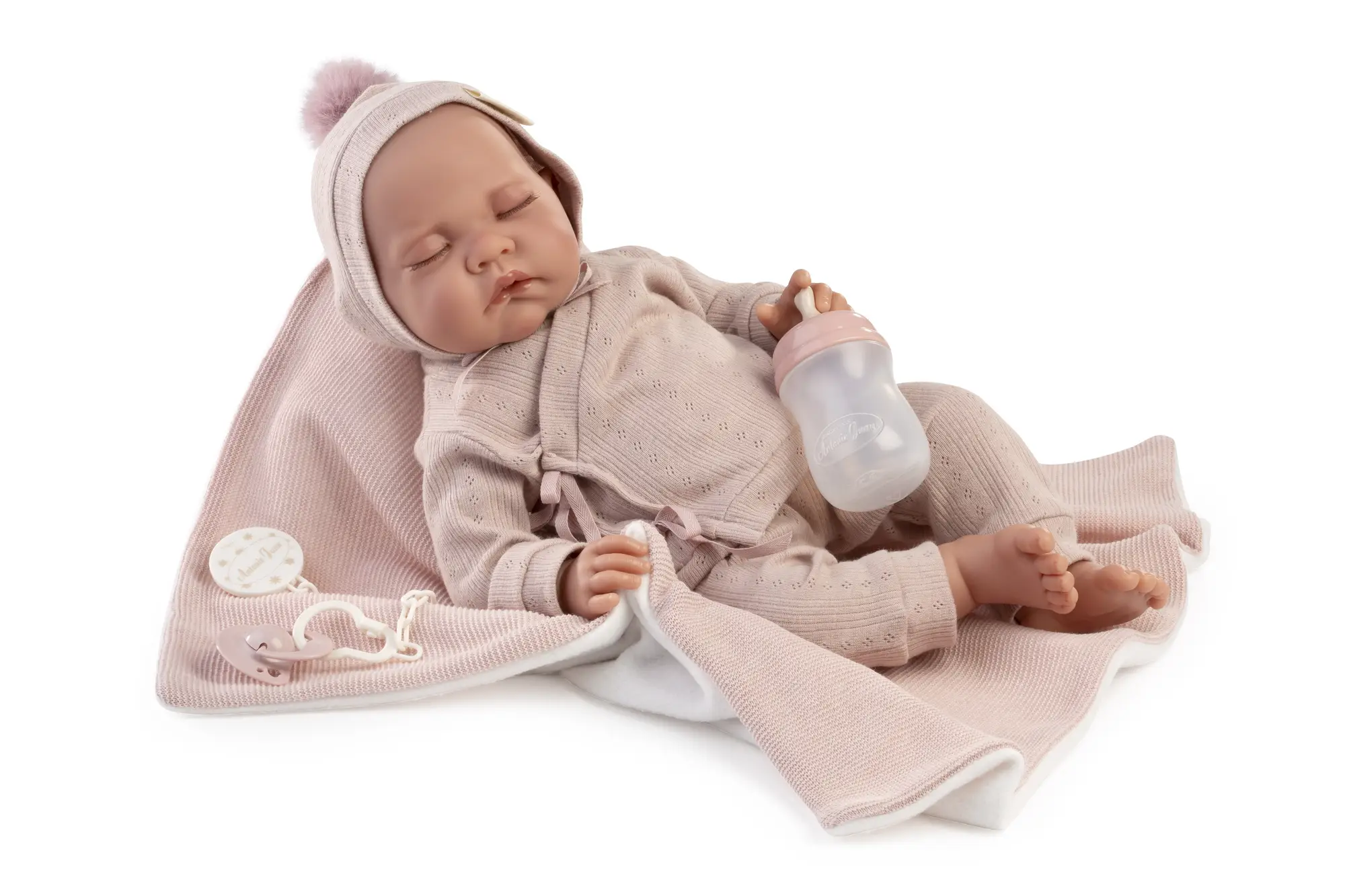 Muñeca Recién Nacida Luna de Antonio Juan dormida sobre una toquilla rosa, con conjunto de punto, gorro con pompón y biberón en la mano.