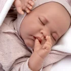 Primer plano de la muñeca Recién Nacida Luna de Antonio Juan dormida sobre un cojín blanco y una toquilla rosa, con una mano junto a la boca.