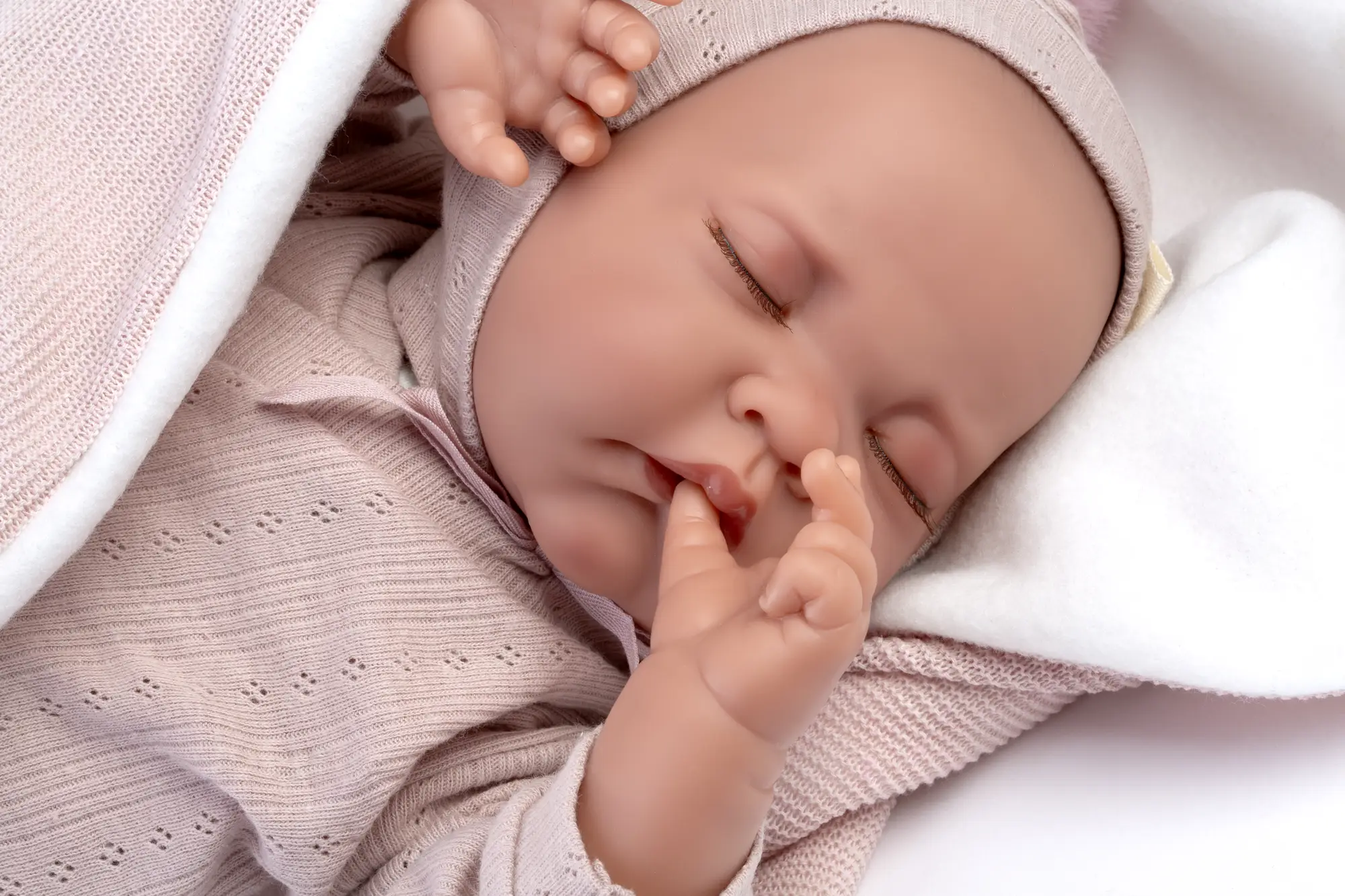 Primer plano de la muñeca Recién Nacida Luna de Antonio Juan dormida sobre un cojín blanco y una toquilla rosa, con una mano junto a la boca.