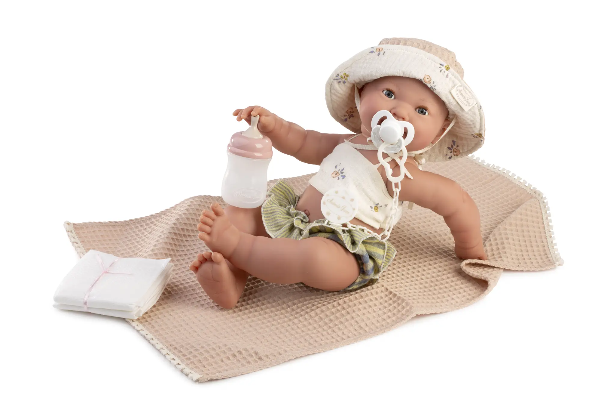 Recién Nacida Mía Playera con biberón y chupete Muñeca Recién Nacida Mía sentada sobre una manta beige con gorro, chupete, biberón y pañales.