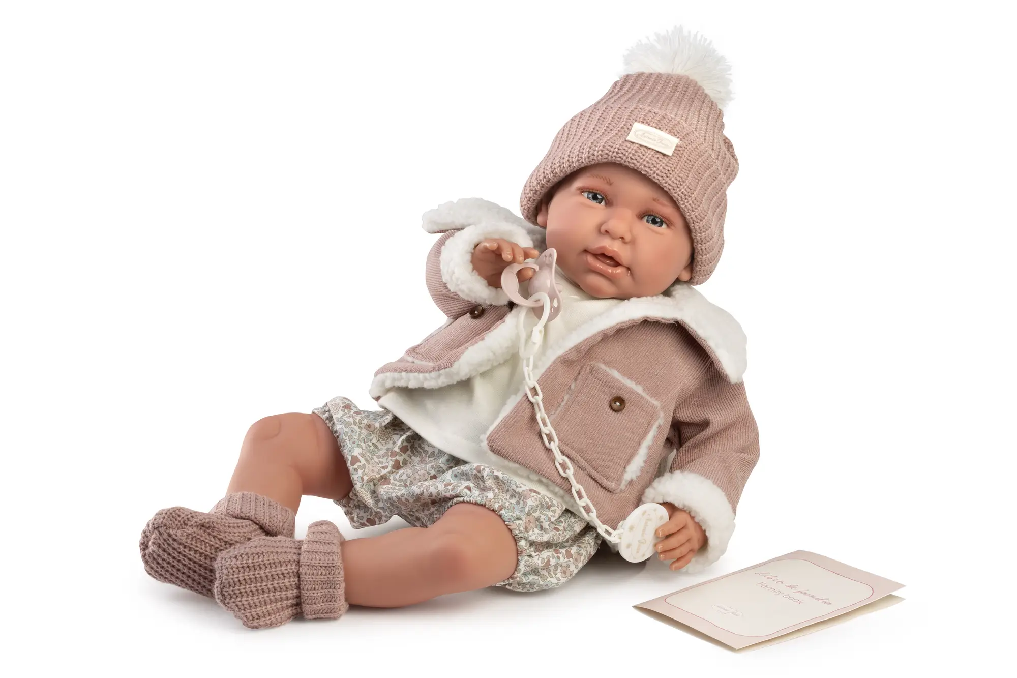 Muñeca Mi Primer Reborn Daniela de Antonio Juan sentada con abrigo rosa, gorro, patucos y chupete sujeto a la mano.
