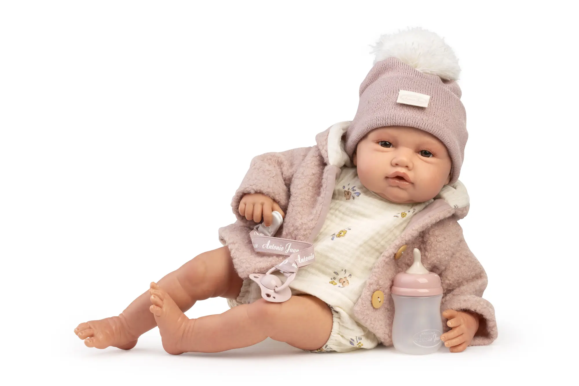 Muñeca Reborn Baby Love Invierno sentada con abrigo y gorro rosa, junto a un biberón y con el chupete en la mano.