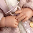 Detalle de las manos y el chupete rosa de Reborn Baby Love Invierno sobre el vestido floral y el abrigo.
