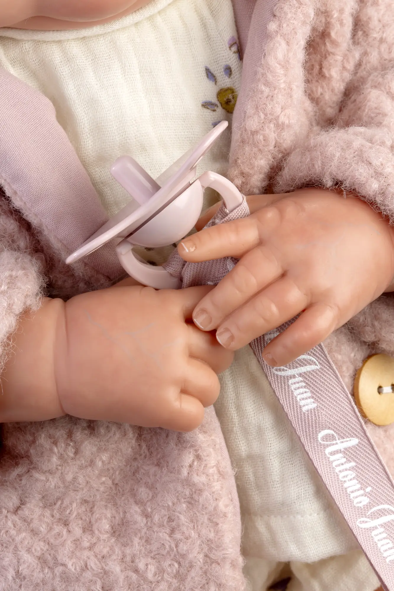 Detalle de las manos y el chupete rosa de Reborn Baby Love Invierno sobre el vestido floral y el abrigo.